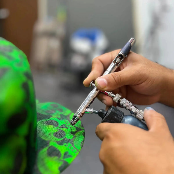 A person’s hands holding an airbrush, applying green and black paint to an object.